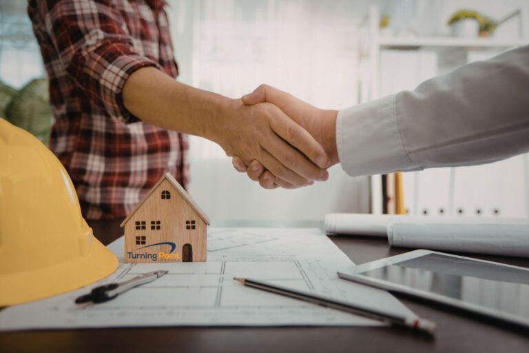 Handshake across architectural plans with a small wooden house model and a yellow hard hat on the desk, symbolizing a construction agreement