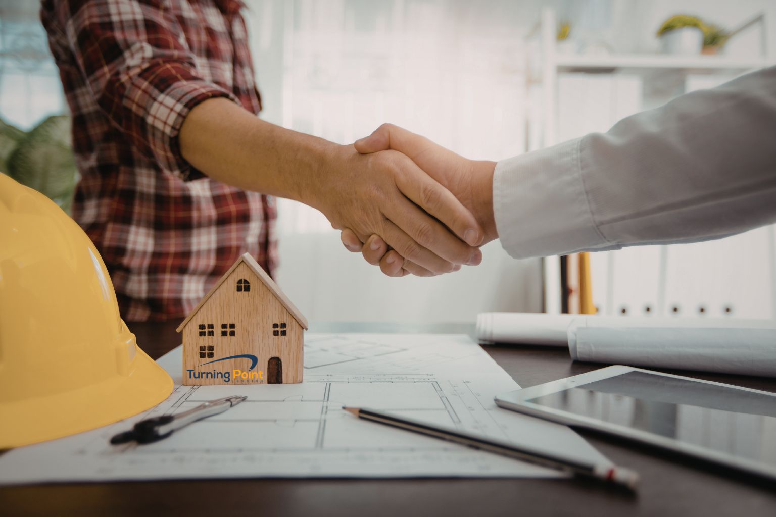 Handshake across architectural plans with a small wooden house model and a yellow hard hat on the desk, symbolizing a construction agreement